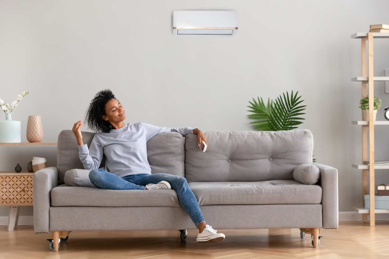 Woman sitting on a gray couch in a living room, looking comfortable while ductless mini-split blows air from top of wall behind her.